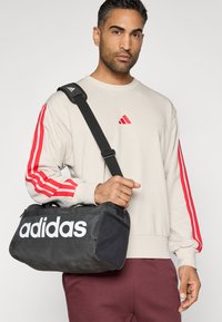 Μαύρη τσάντα duffel με λευκό λογότυπο "adidas", με υφασμάτινη επιφάνεια και πλέγμα στο πλάι, κρατιέται από ένα άτομο που φοράει ελαφρύ φούτερ.
