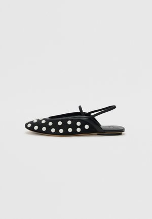 Black flat shoe with a rounded toe, featuring white circular studs across the upper and a slender ankle strap. Smooth texture and minimal design.