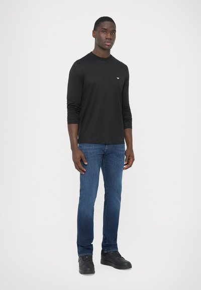 Camiseta negra de manga larga con cuello redondo, combinada con jeans de mezclilla azul y zapatillas negras. Presenta un pequeño logo en la camiseta.