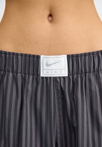 Pantalones cortos de tela a rayas grises y negros con cintura elástica. Presenta una etiqueta del logo de Nike en blanco con un swoosh plateado en la parte delantera.