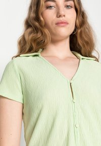 Camisa de manga curta verde claro texturizada com decote em V, fechamento de botão e um sutil efeito amassado. Tecido macio, ajuste descontraído.