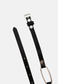 Musta nahkavyö, jossa on hopeinen metallihaara ja pyöreä metallirengas. Tasainen pinta, jossa on vahvistetut saumat ja useita säätöreikiä.