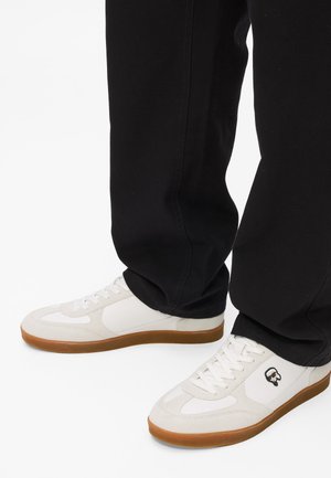 Balti kedai ar zamšas akcentiem, gumijas zoles un mazu logotipa detaļu. Valkāti ar melniem biksēm, demonstrējot gadījuma dizainu un platu formu.