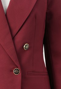 Burgunderfarget blazer med en tettsittende design, teksturert stoff, dobbeltknappet front og gullknappeakenter. Har en sidelomme.