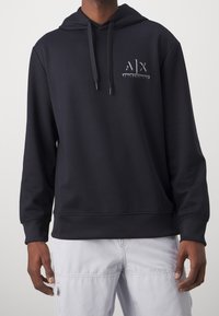 Svart huvtröja med framficka, dragsko i huvan och en texturerad logotyp på bröstet. Tillverkad av mjukt, slätt tyg.