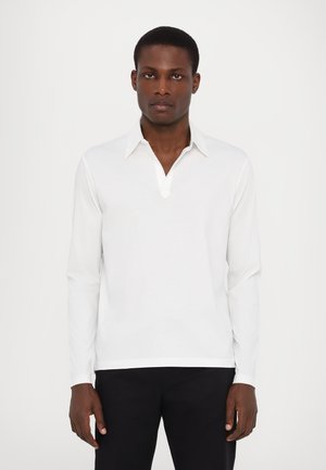 Langarm-Polo aus weißer Baumwolle mit V-Ausschnitt und spitzem Kragen, glatter Textur und geradem Saum, getragen mit schwarzen Hosen.