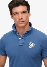 Polo bleu en tissu texturé. Il présente un col, deux boutons au niveau du cou, et un logo crème sur la poitrine.