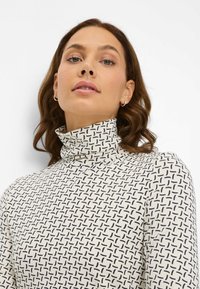Langærmet turtleneck-top i creme med et geometrisk sort mønster. Har tætsiddende krave og glat stoftekstur.