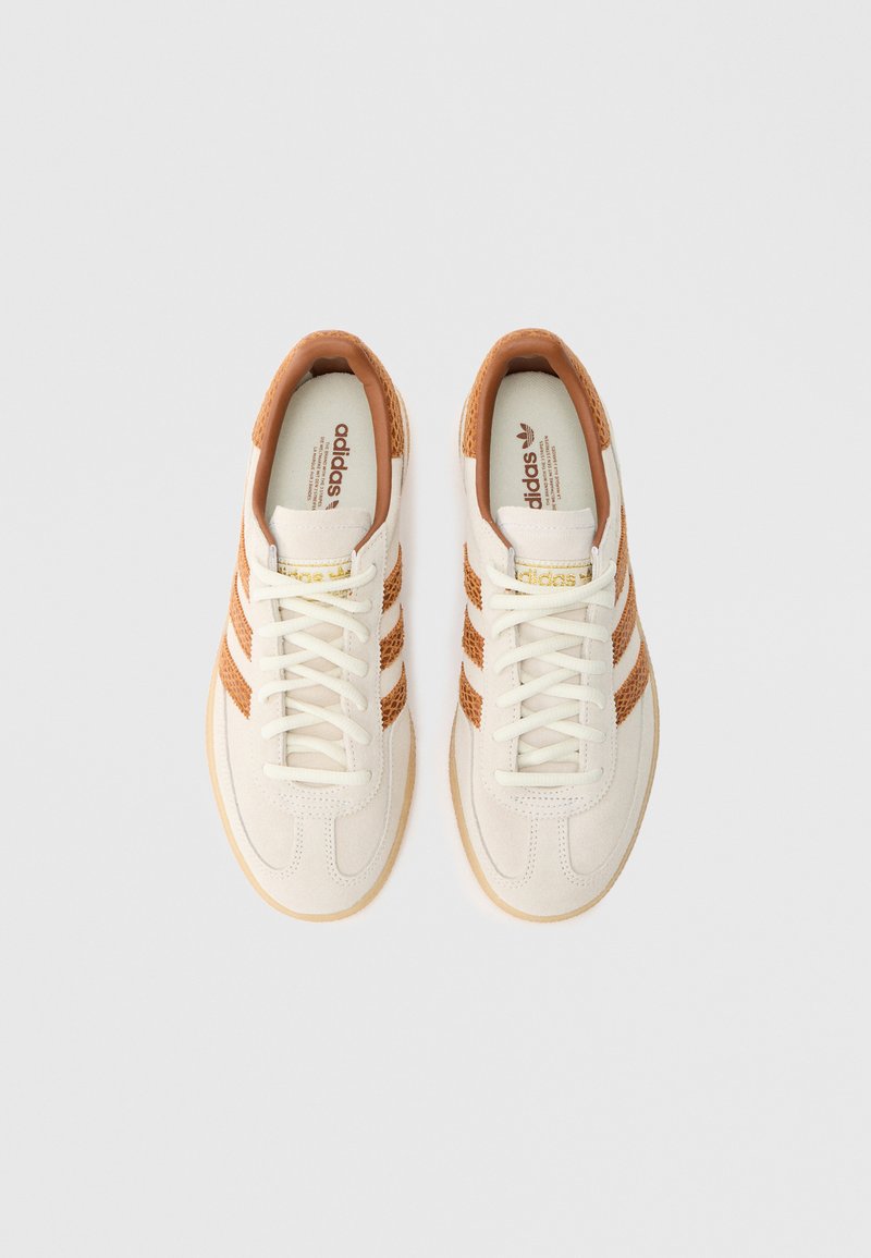 Scarpe da ginnastica bianche con accenti marrone chiaro. Presentano tre strisce marroni, un design testurizzato sui lati e una suola in gomma. I lacci sono bianchi.