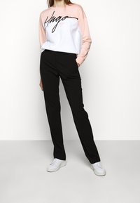 Vrouw gekleed in een roze en witte lange mouw top met zwarte tekst, zwarte broek en witte sneakers, staande met handen in de zakken.