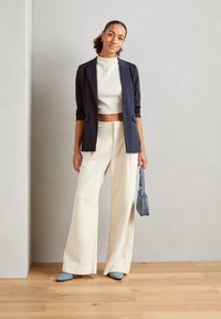 Blazer navy sur un crop top côtelé crème, associé à un pantalon large crème. Tissu texturé, chaussures pointues bleu clair et un petit sac gris.