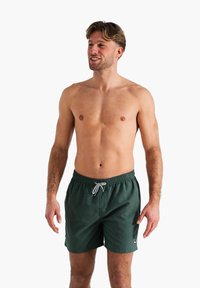 Shorts de bain pour hommes en vert foncé avec une taille élastique, fermeture à cordon de serrage et poches latérales. Tissu lisse, design arrivant au genou.