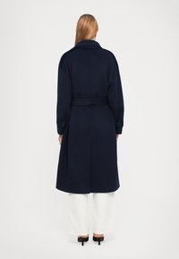 Navy belted coat with a wide collar, long sleeves, and a loose fit, paired with white wide-legged pants and black heels.