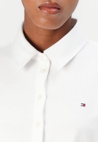 Witte polo met een knoopsluiting bij de kraag, gemaakt van een textuurstof en voorzien van een klein geborduurd logo in blauw, wit en rood op de borst.