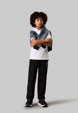 Weißes Polohemd, schwarze Jogginghose mit blauer Druckschrift und schwarze Turnschuhe. Ein navy- und hellblau kariertes Sweatshirt über die Schultern drapiert.