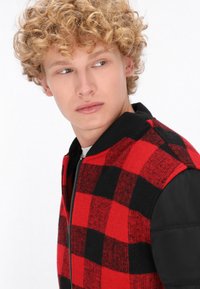Rood-zwart geruit bomberjack met een gebreide kraag en zwarte, gewatteerde mouwen; voorzien van een ritssluiting aan de voorkant en een gestructureerde stof.