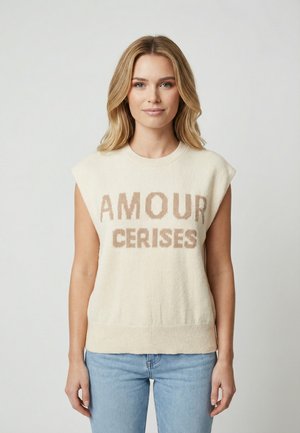 Donna con capelli biondi che indossa un maglione crema senza maniche con la scritta "AMOUR CERISES" e jeans azzurri chiari, in piedi davanti a uno sfondo neutro.