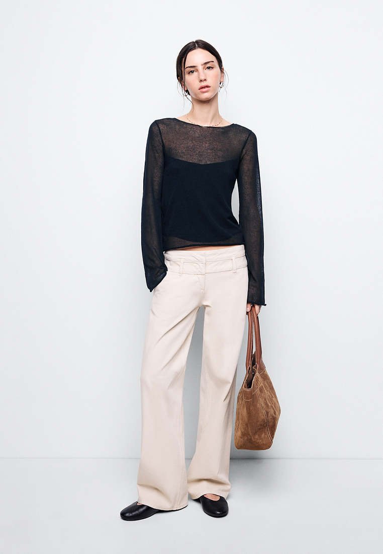 Femme portant un haut noir transparent à manches longues, un pantalon beige à jambes larges, des chaussures noires, tenant un grand sac à main en daim marron, debout devant un fond uni.