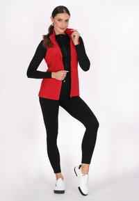 Rote ärmellose Fleece-Weste mit hohem Kragen, Reißverschluss vorne, kombiniert mit einem schwarzen Langarmtop und schwarzen Leggings, vervollständigt durch weiße Sneakers.