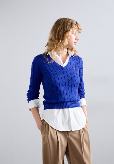 Zils adījuma džemperis ar V-veida kakla izgriezumu, uzsists virs baltas pogu krekla. Tam ir ribotas piedurknes un apmale. Kombinēts ar bēšiem plisētiem biksēm.