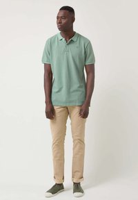 Camisa polo de algodón verde con mangas cortas, combinada con pantalones beige claros y zapatillas verdes. Diseño simple, sin patrones ni acentos visibles.