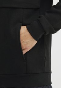 Sudadera negra de tejido suave con un bolsillo frontal y puños acanalados. Presenta una etiqueta en la costura lateral. Primer plano de una mano en el bolsillo.