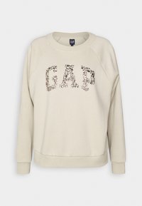 Felpa beige con maniche raglan e scollatura rotonda. Logo "GAP" realizzato con decorazioni metalliche sul davanti. Texture della stoffa morbida.