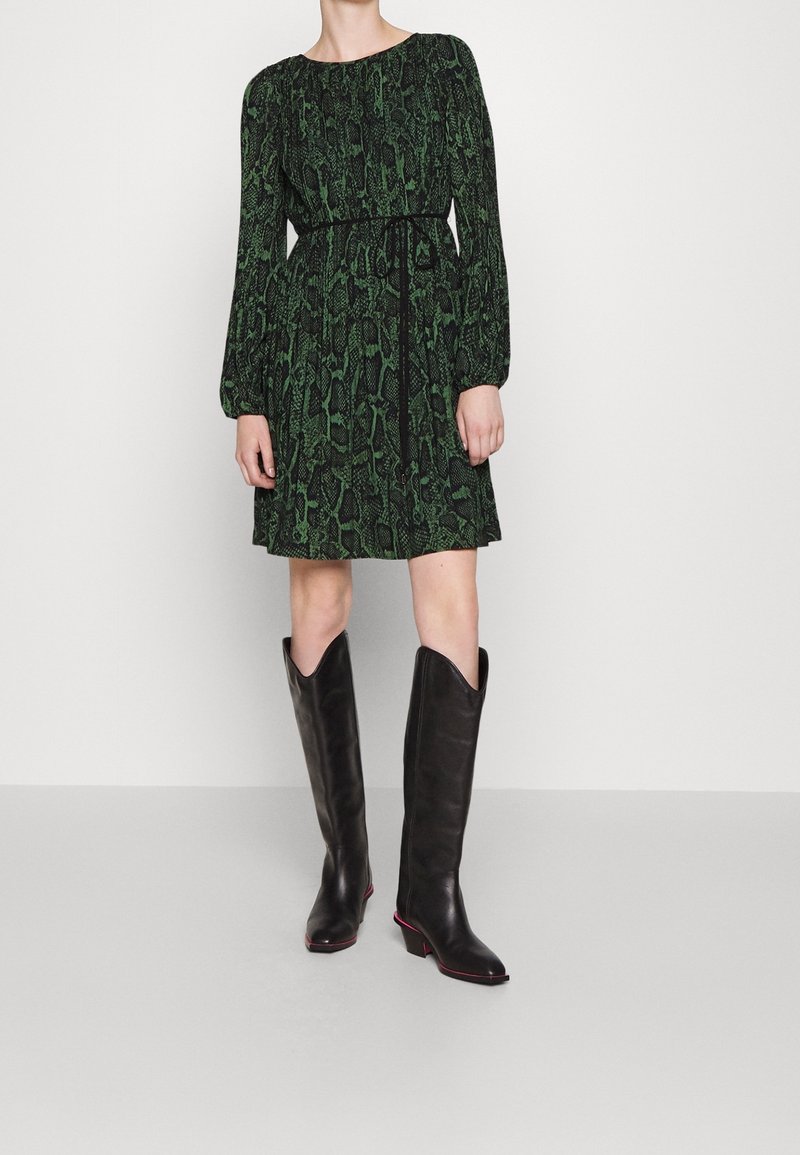 Femme portant une robe à manches longues verte et noire à motifs avec une ceinture noire et des bottes en cuir noires montant jusqu'aux genoux.