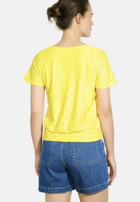 Dzeltena īsām piedurknēm blūze ar ķīļveida rakstu, apaļu kakla izgriezumu un nedaudz pieguļošu jostas vietu. Apvienota ar ziliem džinsu šortiem.