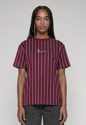 Jeune personne aux longs cheveux tressés portant un t-shirt à manches courtes bordeaux avec des rayures blanches verticales et un pantalon noir, debout devant un fond uni.