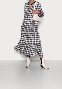 Dlouhé šaty s černobílým kostkovaným vzorem a vrstvenými volány, s límečkem, zapínáním na knoflíky a kombinované s bílými kotníkovými botami a pletenou taškou.