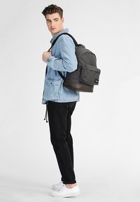 Mochila de tela gris con parte inferior marrón, que cuenta con correas acolchadas y un bolsillo frontal; la lleva puesta una persona con una chaqueta de mezclilla azul clara y pantalones negros.