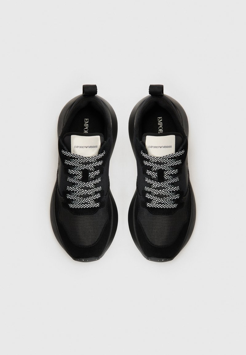 Zapatillas deportivas negras con parte superior de malla, acentos texturizados, cordones blancos en contraste y diseño de puntera redondeada. Etiqueta de marca visible en el interior.