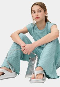 Turquoise textuuroutfit met golvend patroon, geschulpte zoom, gecombineerd met zilveren platte sandalen en een kleine metalen schoudertas.