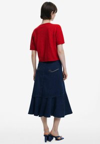 Top corto a maniche corte lavorato a maglia rossa abbinato a una gonna svasata in denim scuro con un design a strati e tasche posteriori. Indossato con sandali con tacco blu.
