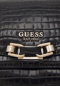 Schwarze Handtasche aus Kunstleder mit Krokodillederstruktur, goldenen Akzenten und einem auffälligen GUESS-Logo mit "EST 1981" darunter.