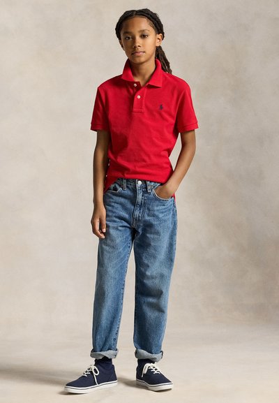 Kind met gevlochten haar, gekleed in een rood poloshirt, blauwe opgerolde spijkerbroek, donkerblauwe sneakers, staand met één hand in de zak tegen een neutrale achtergrond.