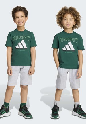 To smilende børn står side om side iført grønne Minecraft grafik t-shirts, lysegrå shorts og sorte-grønne sneakers.