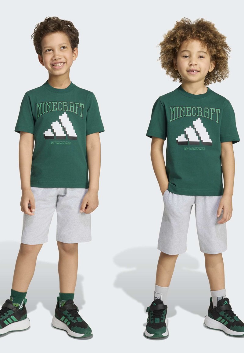 Deux enfants souriants se tiennent côte à côte, portant des t-shirts verts avec un motif Minecraft, des shorts gris clair et des baskets noir-vert.