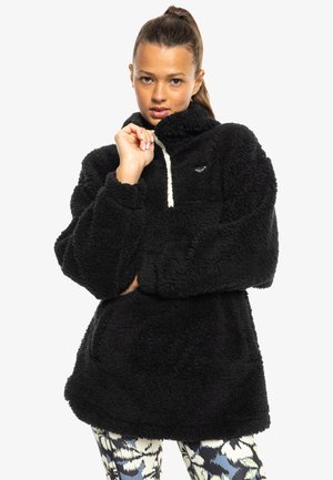 Black fleece pullover with a high collar, cream drawstring, and a small logo. Features wide sleeves and a relaxed-fit design.
