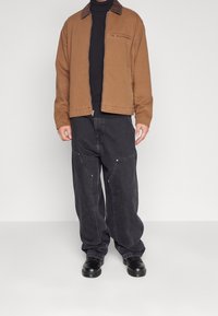 Brun mellemvægtig jakke med en lynlås-lomme, parret med løst siddende sorte jeans. Sorte sko fuldendte outfittet.