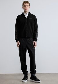 Casaco desportivo preto com fecho de correr, detalhes em riscas cinzentas, combinado com calças de treino pretas a condizer e sapatilhas pretas. Apresenta um design simples e direto.