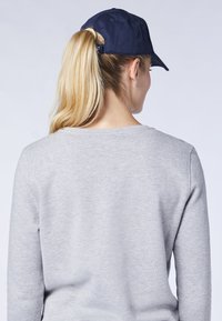 Marineblaue Baseballmütze mit gebogenem Schild, rückwärts über langen, glatten blonden Haaren getragen, kombiniert mit einem hellgrauen Pullover.