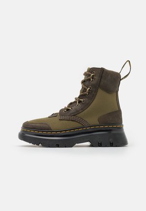 Wysokie buty trekkingowe w kolorze oliwkowo-zielonym i brązowym z grubą czarną podeszwą, żółtym przeszyciem i sznurowaniem, widoczne z boku.