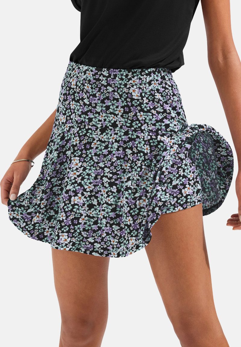 Cvjetna suknja s crnom pozadinom, koja sadrži mala mente zelena i ljubičasta cvijeća. Elastični pojas, lepršavi dizajn dužine do sredine bedra.