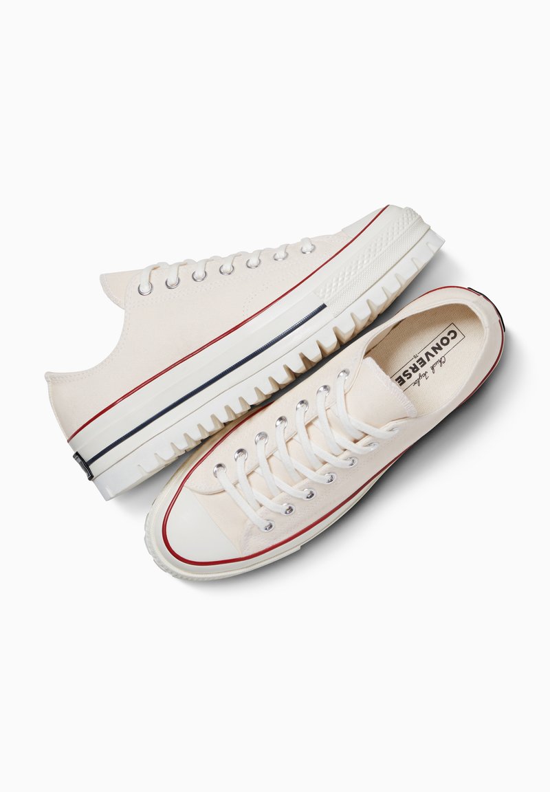 Weiße Low-Top-Sneaker aus Canvas mit weißen Schnürsenkeln, rotem Streifen an der Sohle, schwarzem Streifen an der Zwischensohle und sichtbarer markierter Innensohle bei einem Schuh.