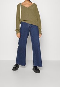 Olivgrüner V-Ausschnitt-Pullover aus weichem Material, kombiniert mit weit geschnittenen, dunklen Denim-Jeans. Das Modell trägt schwarze Plateauschuhe. Ein einfacher Taschenriemen ist sichtbar.