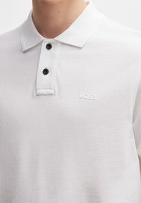 Biała koszulka polo z teksturowanym wzorem, z klasycznym kołnierzykiem, dwoma czarnymi guzikami i małym, wytłoczonym logo "BOSS" na piersi.