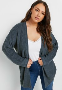 Lungo cardigan grigio a maglia con apertura frontale, texture a coste, maniche ampie e vestibilità casual, abbinato a un top bianco con bordature in pizzo.