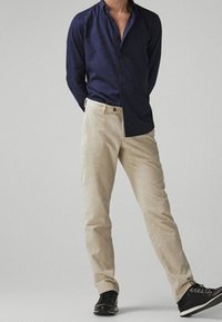 Man staat met zijn handen achter zijn rug, gekleed in een marineblauw overhemd met knopen, beigere broek en zwarte schoenen, op een effen achtergrond.